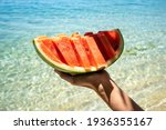 Hand holding slice of watermelon on the beach. Sea and blue sky background. Summer mood. 2021
