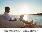 Senior man working on his laptop lying on deck chair on the beach during sunset