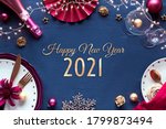 Happy New Year 2021 gilded text in frame made from Silvester party table setup. Vine, glasses, plates, fork, knife. Xmas decorations and garland. Flat lay in golden and red on classic blue textile.