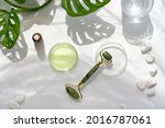 Moisturizer, green jade face roller with exotic leaves. Monstera Adansonii leaves. Sunshine, long shadows. Minimal flat lay on off white background. Facial massage with self made cosmetics.