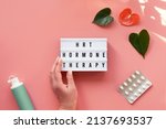 Small photo of Text HRT Replacement Therapy on light box in hand. Menopause, hormone therapy concept. Estrogen replacement therapy awareness design. Pink background with exotic leaves, flowers, pills, estrogene gel.