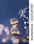 Small photo of End of the year 2022, Silverster, Happy New Year 2023. Hourglass with numbers 2022 and 2023 and light garland on dark blue background. Hourglass is also known as sandglass, sand glass, sand timer or