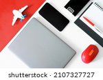 Silver laptop, computer mouse, fountain pen, diary, power bank, mobile phone and toy plane on a red and white table. Concept of preparing for journey, choosing and buying air tickets online. Daylight