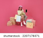 Happy Asian teen woman sitting on sofa holding shopping bags and smartphone isolated on pink background, Shopper or shopaholic concept.