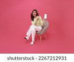 Happy young Asian woman showing empty screen mobile phone. While her sitting on chair isolated on pink studio background.