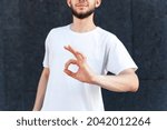 Small photo of Gesture, emotion, expression and people concept - Close-up of the okie sign by the hands of a European bearded man in a white T-shirt outdoors on a black background with copy space