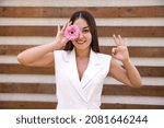 Small photo of blossoms, emotions, gesture and people concept - portrait of an oriental girl in white clothing with a flower by the eye showing an okie sign with hands on a brown ribbed background with copy space