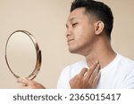 Small photo of Expression of unhappy asian man looking in mirror, feeling stressed of sensitive skin or acne breakout, thinking of cosmetology treatment. Depressed young guy dissatisfied with skin condition.