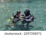 Small photo of Diving with aqualung lesson, instructor giving scuba diving lessons to a kid. Trainer teaching a little boy to dive. Two scuba divers dressed in diving suit is preparing to dive in shallow sea.