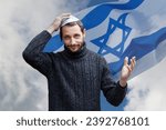 Small photo of Religious jew man with smile face, holding a kippah, hand to his head against the background of the Israeli flag, beautiful sky. Handsome smiling bearded Jewish holding yarmulke for wind over his head