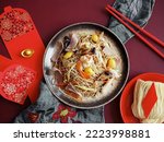 Small photo of Chinese Lunar New Year lucky food longevity noodle or stir fried noodles served on red background with red envelopes, chopsticks, ancient gold bullion nugget and uncooked dried noodles. (top view)