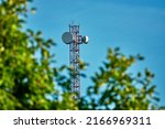 Mast with cell phone antennas against blue sky. Tree with green leaves.