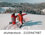 Two girls with snowboards dressed in red snowboarding jackets standing on the mountain looking to a good line, beautiful view in sunny day, bleu sky