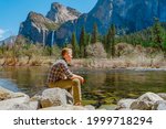 A young man sits on the bank of a river and enjoys the view of a mountain waterfall in Yosemite National Park
