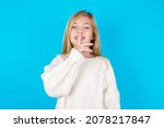 Small photo of Smiling caucasian little kid girl wearing white knitted sweater over blue background makes shush gesture, holds fore finger over lips hides secret. Be mute, please.