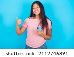 Small photo of Portrait of Young beautiful woman wearing pink T-shirt against blue background using and texting with smartphone happy with big smile doing ok sign, thumb up with fingers, excellent sign