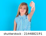 Small photo of Discontent little kid girl with glasses wearing plaid shirt over blue background shows disapproval sign, keeps thumb down, expresses dislike, frowns face in discontent. Negative feelings.