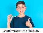 caucasian kid boy wearing blue T-shirt over blue background holding an invisible braces aligner and rising thumb up, recommending this new treatment. Dental healthcare concept.