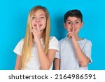 Small photo of Smiling two kids boy and girl standing over blue background makes shush gesture, holds fore finger over lips hides secret. Be mute, please.