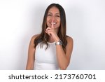 Small photo of Smiling young beautiful caucasian woman wearing white top over white background makes shush gesture, holds fore finger over lips hides secret. Be mute, please.