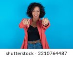 Close-up portrait of surprised young beautiful woman with curly short hair wearing red overshirt over blue wall pointing with two fingers to the camera saying: I choose you!, looking up with open mout