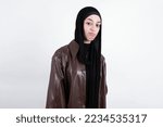 Small photo of beautiful muslim woman wearing hijab and brown leather jacket over white background with snobbish expression curving lips and raising eyebrows, looking with doubtful and skeptical expression