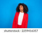Small photo of young businesswoman with afro hairstyle wearing red over blue wall with snobbish expression curving lips and raising eyebrows, looking with doubtful and skeptical expression, suspect and doubt.