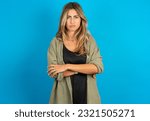 Small photo of Gloomy dissatisfied young beautiful blonde woman wearing overshirt looks with miserable expression at camera from under forehead, makes unhappy grimace