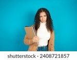 Small photo of Portrait of dissatisfied Beautiful student girl holding books wearing formal clothes smirks face, purses lips and looks with annoyance at camera, discontent hearing something unpleasant