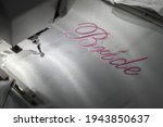 Machine embroider the inscription bride on a white satin robe.
Inscription bride with pink thread. Wedding day preparation. Elegant bridal lingerie.White accessories for the bride.Glamorous style.
