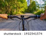 Human hands holding bicycle handlebar against landscape with country road. Man travelling by bicycle in northern forest in summer evening. Weekend activities, healthy lifestyle, adventure concept. POV