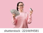 Small photo of Bank card. Portrait of young attractive caucasian woman wearing glasses shows a bank card and a fan of dollar bills. Pink background. The concept of money remittance and stock market.