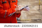 Small photo of Action of a safety officer in full PPE coverall is writing note on paper document during perform safety audit at construction worksite. Industrial expert working scene. Selective focus.