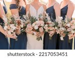Bride and bridesmaids holding bouquets. Bridesmaids are wearing navy blue dresses. Bouquet has roses and wildflowers. 