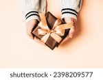 Girl's hands in white sweater holding brown gift box with golden bow on light peachy background. Christmas, Valentines day, Black friday sale concept. Top view. Peach Fuzz color of the year 2024