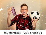 Small photo of Ecstatic fan, young man holding ticket for soccer match and soccer ball. Confetti in the back. Man winning ticket as prize