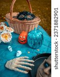 Small photo of halloween decorations on picnic blanket with picnic basket with black guise. head on plate with skeleton hands, skull decorations, pumpkin, flowers, seasonal