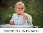Small photo of happy smiling one year old baby boy in chic festive summer clothes looking flirty into the camera outdoor in the garden with magical, idyllic flowery green grass in the background