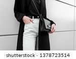 Woman in a trendy T-shirt in a black long fashionable coat in stylish white jeans with a black leather elegant bag. Trendy elegant casual outfit. Details of everyday look. Street fashion. Close-up.