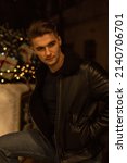 Small photo of Fashionable handsome brutish man in a black leather jacket stands outside with Christmas decorations and lights at night