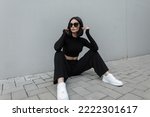 Fashionable young urban woman with black trendy sunglasses in fashion black stylish sports outfit with white sneakers sits on the street near a modern gray building