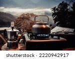 Small photo of Outdoor kitchen equipment camp fire and brewing tea pot moka coffee drip cup,with wooden table camping gas stove set in nature outdoor,mountain background,concept camp and travel,vintage tone color