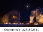 Small photo of Jabal AlFil - Elephant Rock in evening, landscape illuminated, text Alula and Arabic translation projected to stone wall seats for people set up on the ground, crescent moon - composite image - above