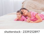 Small photo of Asian happy cute big sister playing with adorable newborn baby girl in morning after waking up. elder sister teasing toddler while lying on bed together. Family relationships concept.