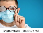 Man wiping foggy glasses caused by wearing disposable mask on blue background, space for text. Protective measure during coronavirus pandemic