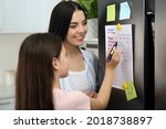 Young woman with her daughter checking to do list on fridge in kitchen