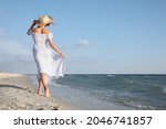 Woman in dress with straw hat walking by sea on sunny day, back view