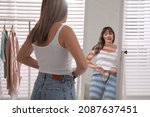 Small photo of Young woman trying to put on tight jeans near mirror at home