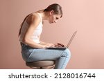 Small photo of Woman with bad posture using laptop while sitting on stool against pale pink background