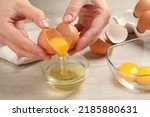 Woman separating egg yolk from white over glass bowl at wooden table, closeup
